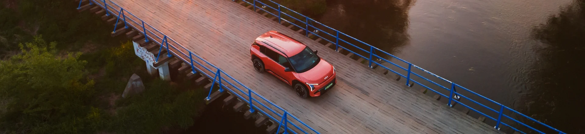 Baner promocyjny Kia EV3 w kolorze Terra Cotta – kompaktowy, elektryczny crossover SUV zaparkowany na środku mostu nad rzeką, widok ¾ z góry