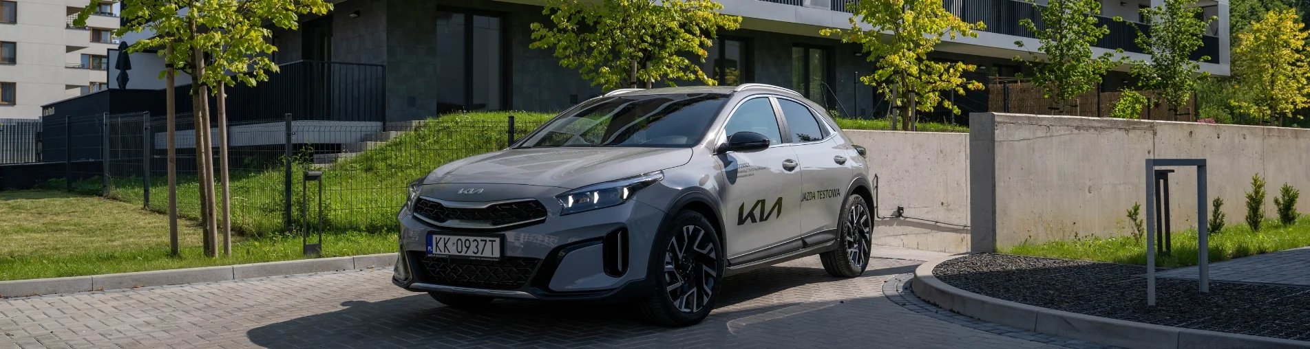 Baner Kia XCeed w kolorze Wolf Grey – srebrnoszary crossover‑SUV zaparkowany pod blokami mieszkalnymi, widok ¾ z przodu